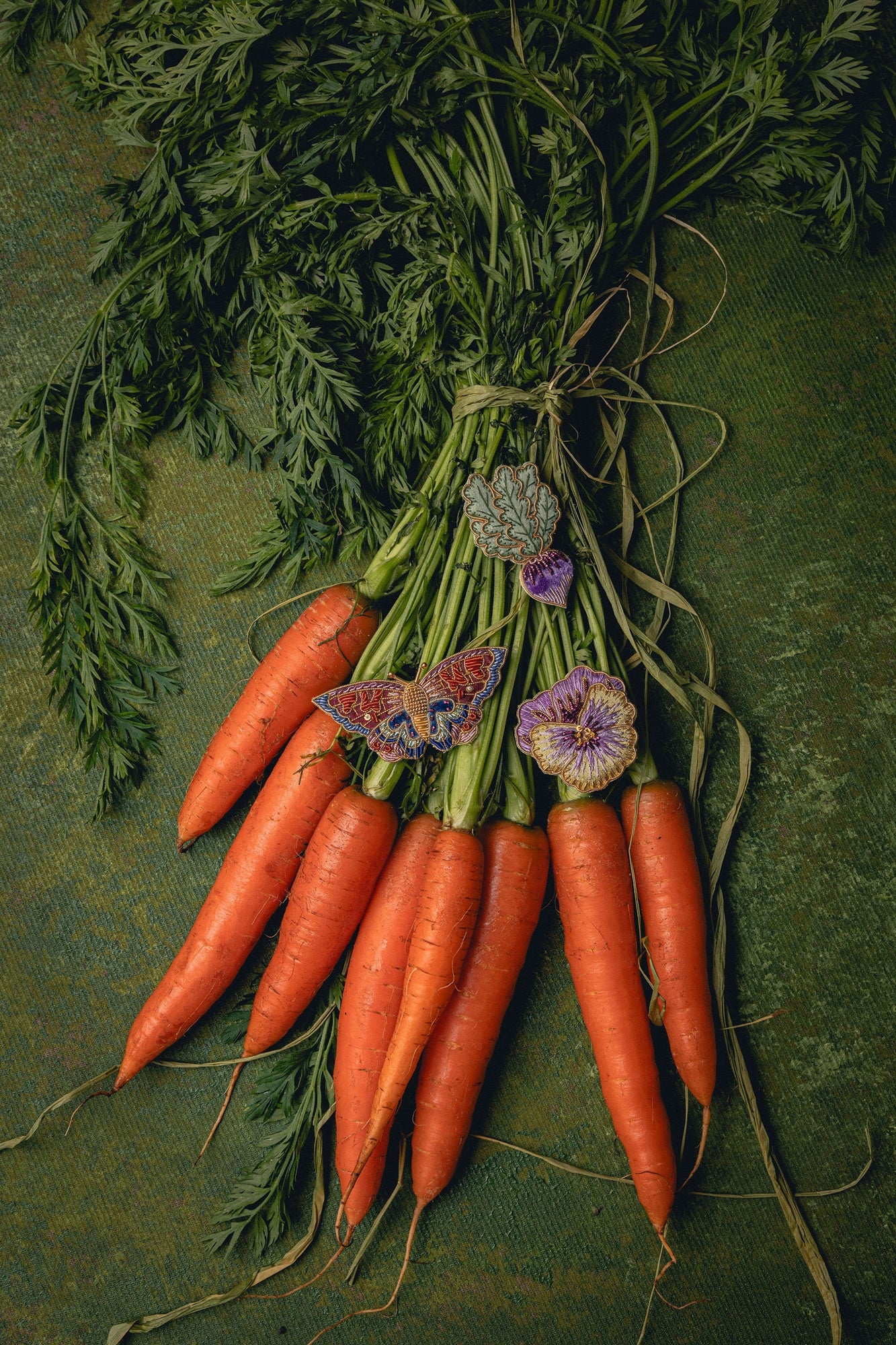 How Does Your Garden Grow? Collection of Hand Embroidered Jewellery - Bunch of carrots with green leaves on a textured green background with brooches displayed on top