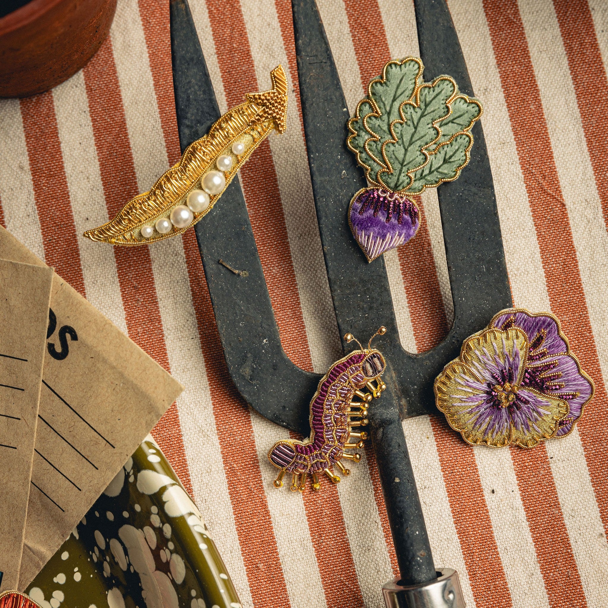 How Does Your Garden Grow? Collection of Hand Embroidered Jewellery - Decorative brooches, displayed on a gardening hand fork on a striped fabric background