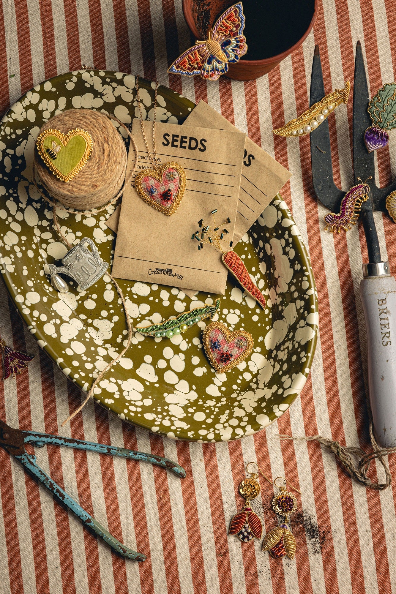 How Does Your Garden Grow? Collection of Hand Embroidered Jewellery - Decorative plate with embroidered jewellery, seeds packets and gardening tools on a striped background