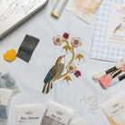 The Bird & Blossoms Kit - Embroidery kit with a completed bird and floral design on fabric, surrounded by materials